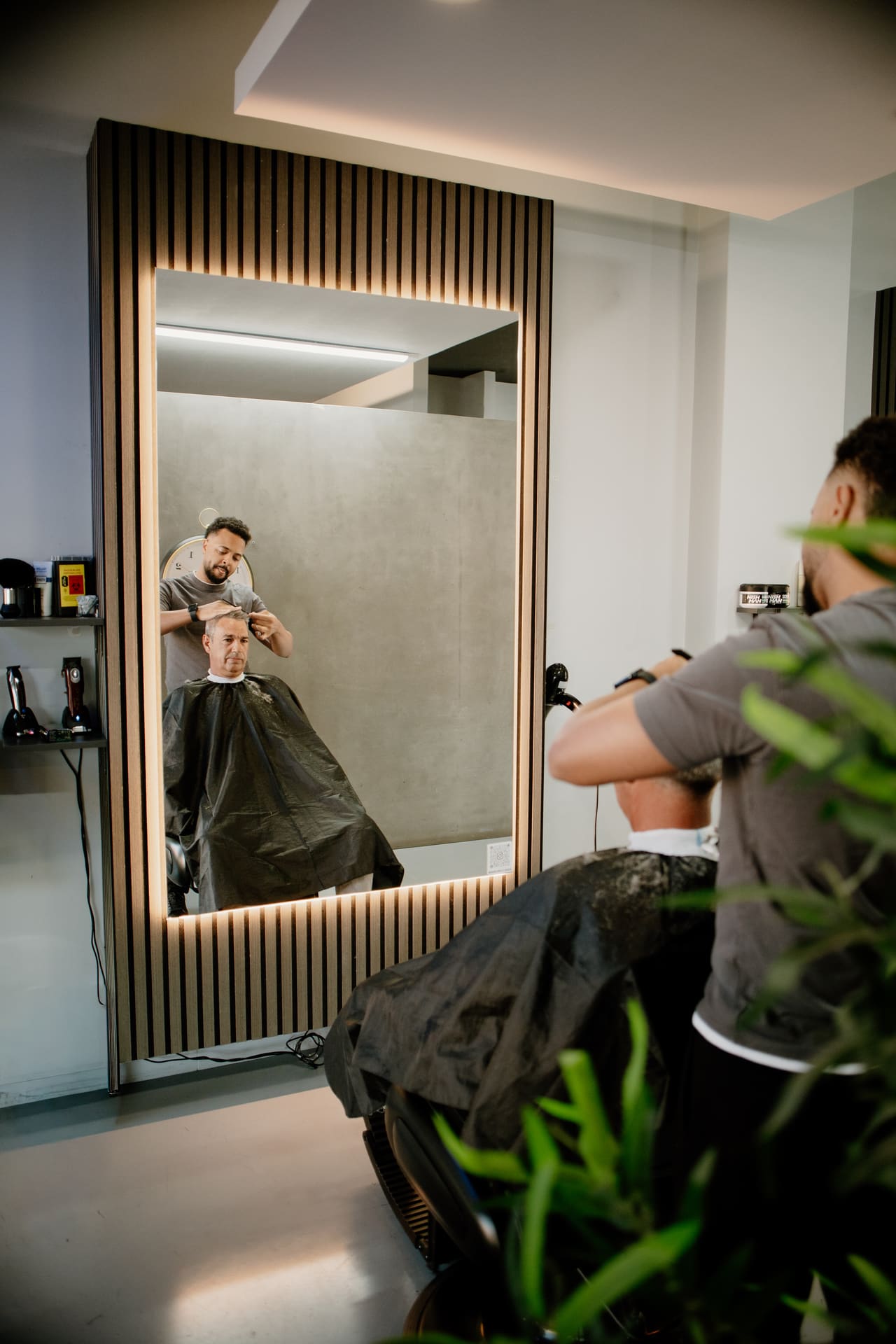 Reflexo no espelho de um barbeiro a cortar o cabelo de um cliente, com plantas em primeiro plano