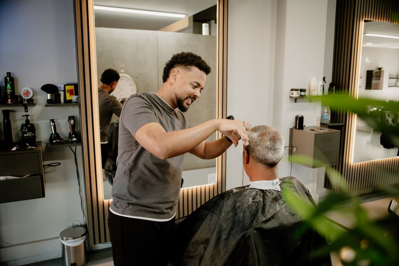 Barbeiro a sorrir enquanto acerta o corte de um cliente, junto ao espelho iluminado
